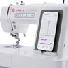 White Singer Smart Heavy Duty sewing machine with a smartphone app screen mounted on a dark green textured panel, shown on a clean white background.
