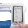 A close-up of a white Singer smart heavy duty sewing machine hd9120 with a smartphone mounted in a green holder. A finger is touching the phone screen, which displays a thread selector list, and blue fabric is visible on the sewing needle plate.