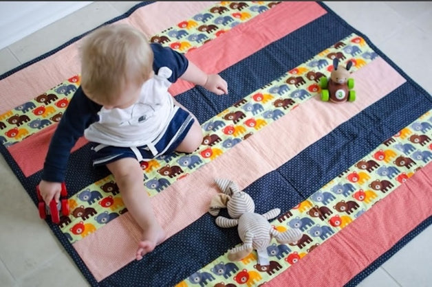 Toddler playing on a colorful striped quilt featuring bear patterns in pink, blue, and yellow, with toys including a toy car and a stuffed bunny, on a tiled floor.